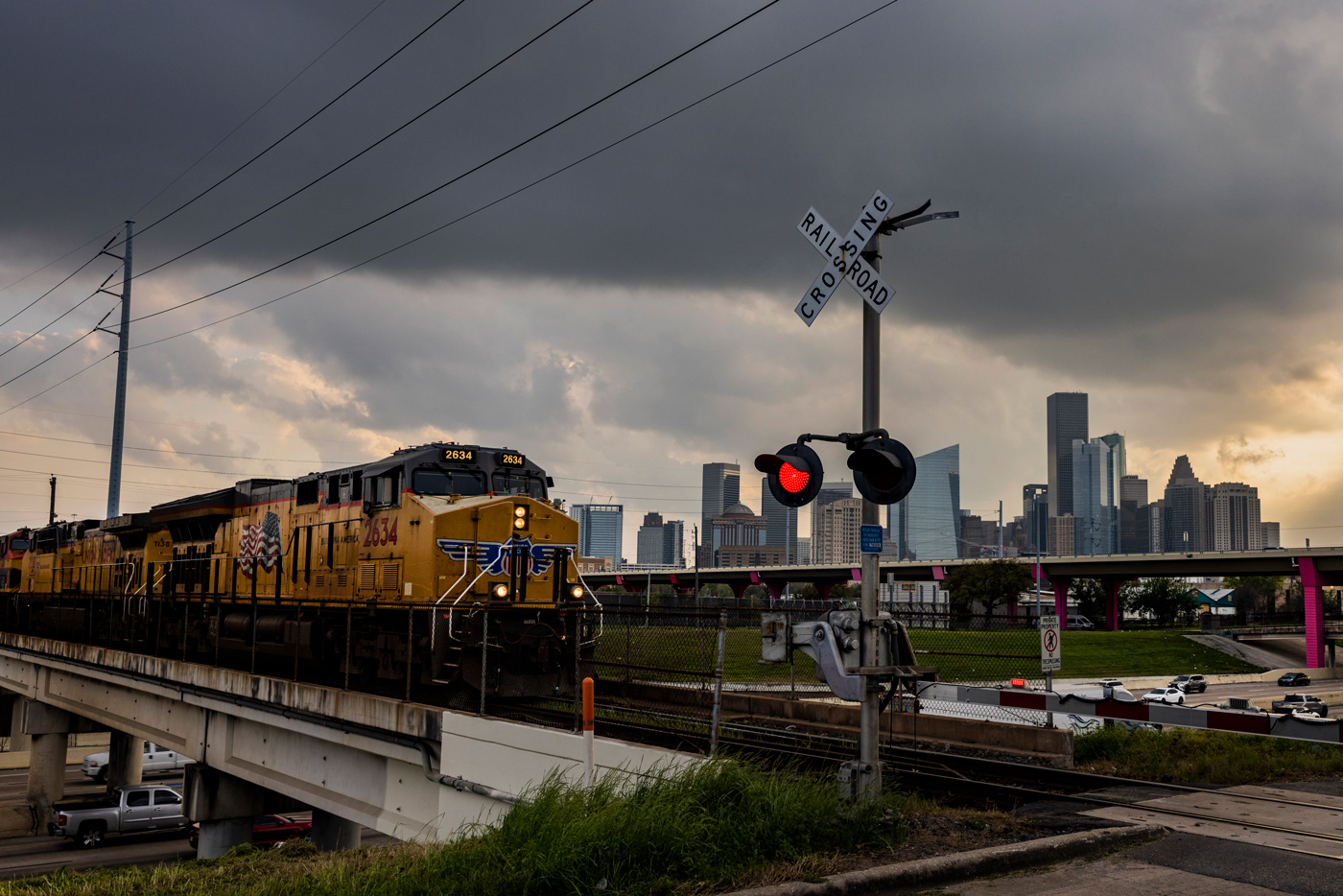 A train approaches a railroad crossing with the skyline of downtown Houston on the background.